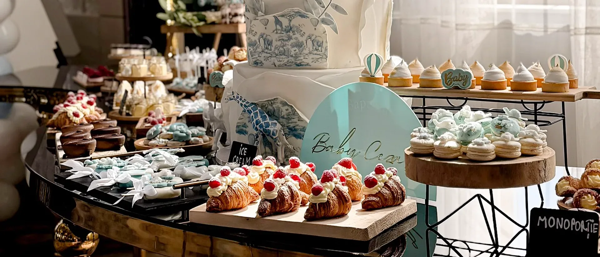 Tort etajat alb cu bleu pentru botez, alături de croissante cu frișcă și zmeură, macarons și prăjituri asortate pe masa de candy bar